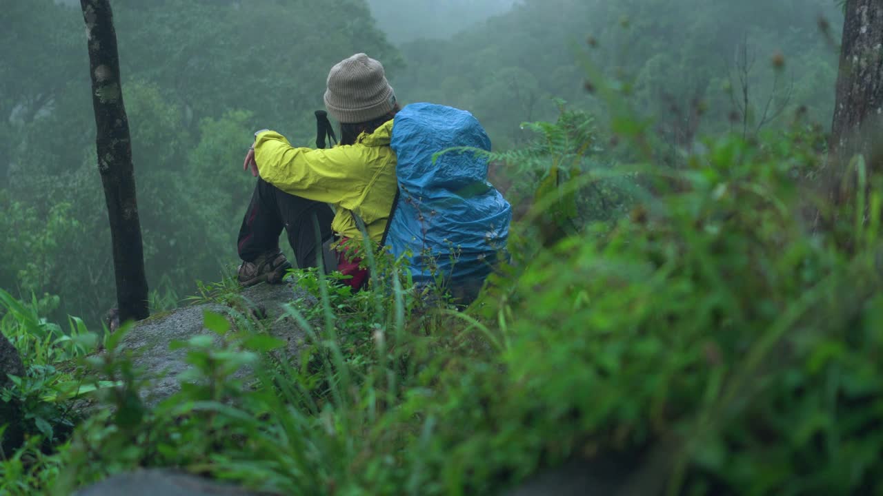 亚洲探险家，冒险家，女性背包客在恶劣的天气下独自徒步旅行的热带森林，在泰国的雨季，雾覆盖了所有的山峰，包括下雨，寒风。坐在岩石上休息，几乎是晚上视频素材