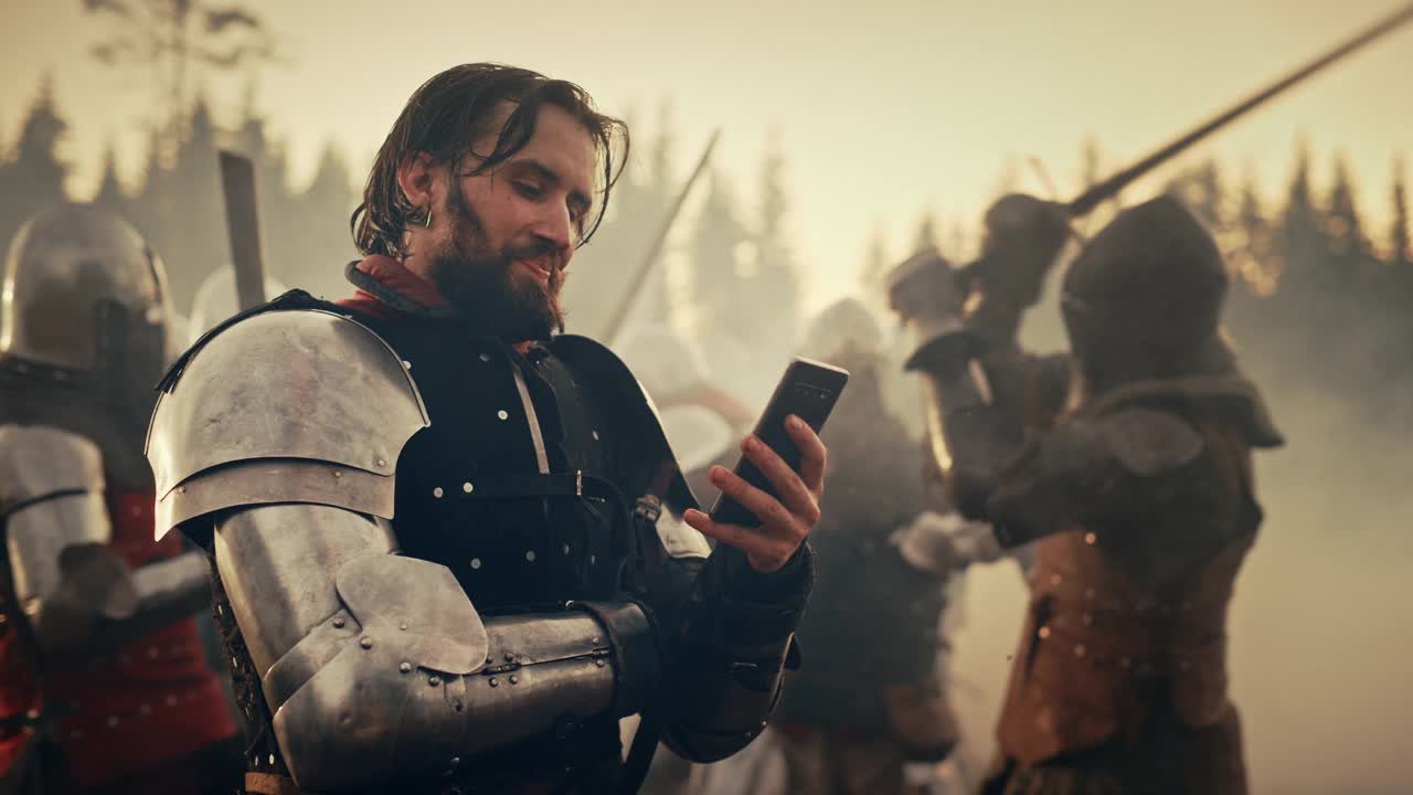 英俊的中世纪骑士在战场上微笑着使用智能手机。有趣的概念:成功的装甲战士网上订购，赌博，投资，做电子商务。战争是激烈的视频下载