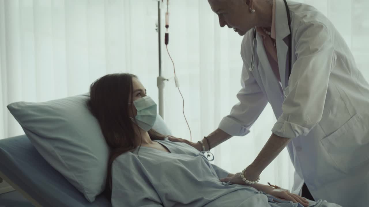 一位女病人躺在病床上，与她的资深医生讨论处方药物的选择。医生回答病人的问题。他们戴着防护口罩。视频素材