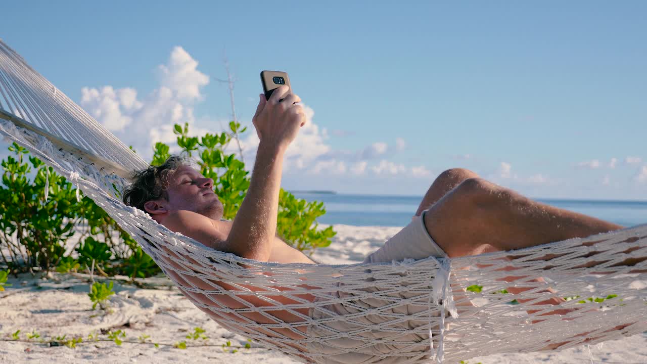 一名男子躺在沙滩上两棵棕榈树之间的吊床上打电话视频下载