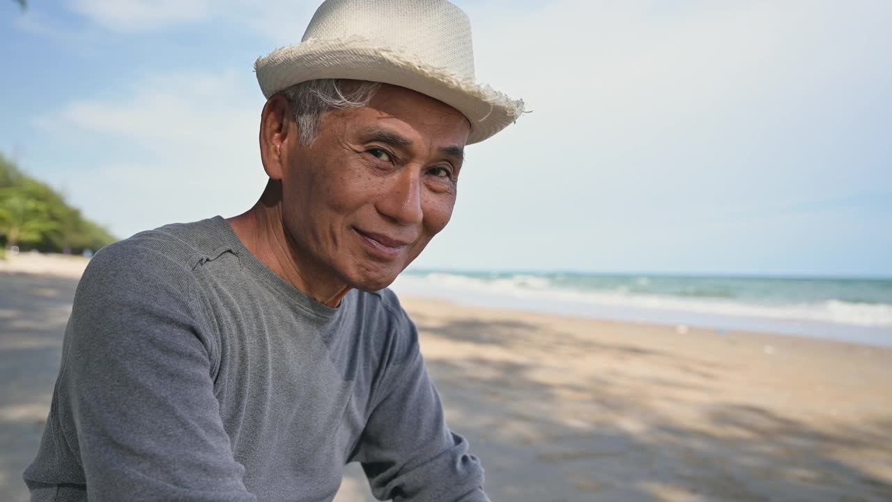 亚洲老人戴着帽子坐在沙滩上微笑着用手势打电话聊天。视频下载