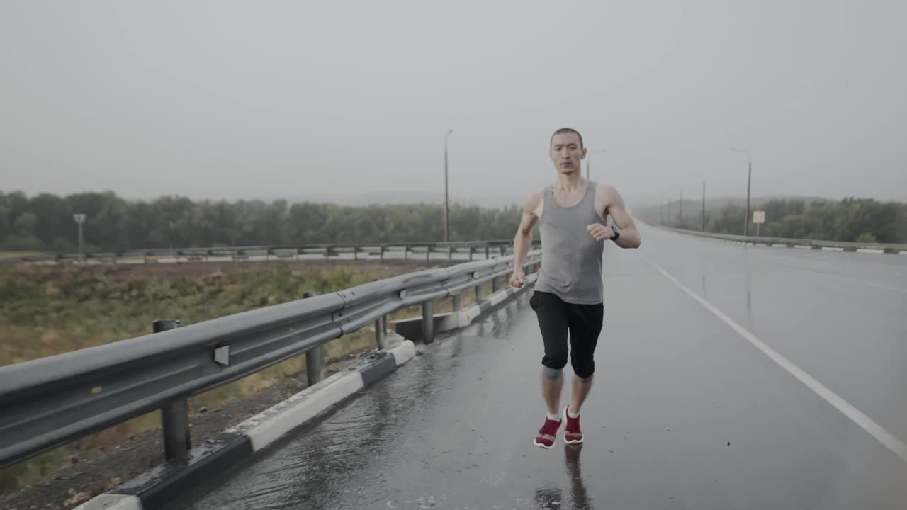 亚洲模样的运动员，体格健壮，穿着运动鞋沿着潮湿的沥青公路穿过水坑跑步，正面看雨暴风雨的天气。抱负和速度的概念视频素材