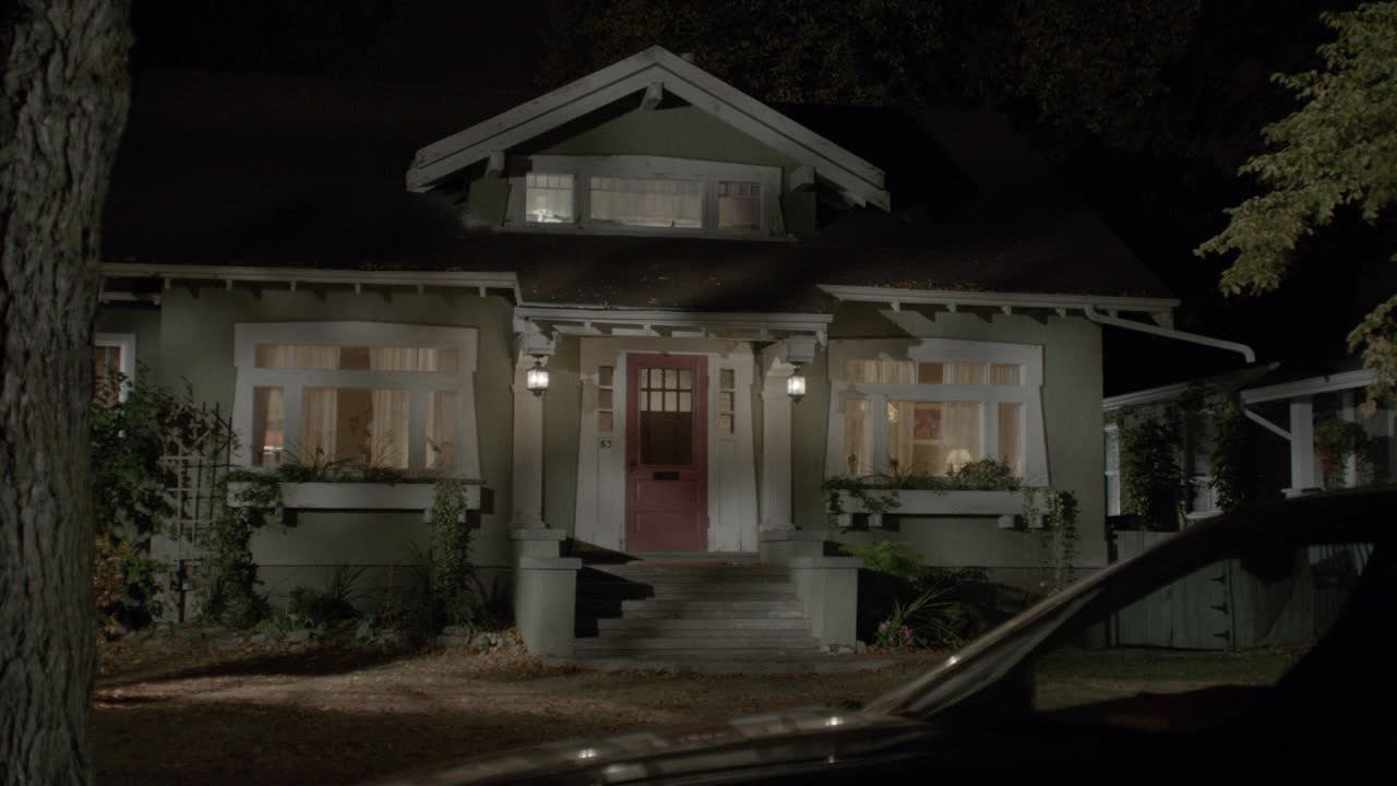 中等角度的绿色加州工匠风格的房子，白色装饰和红色前门。房子里面亮着灯。部分可见的汽车停在街道上。可以是中产阶级社区、郊区或住宅区。匹配dx / nx 4318-001, 4318-0视频下载