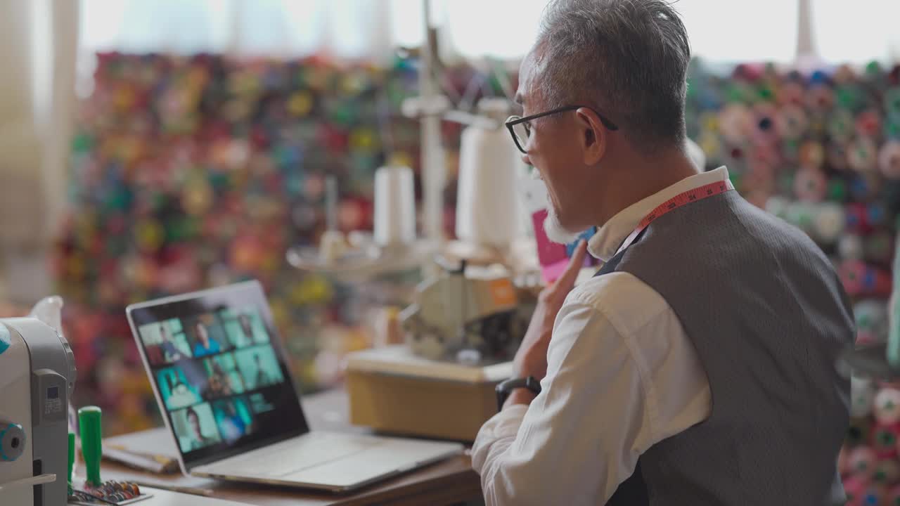 一名亚洲华裔老人正在展示布料样本，视频呼叫一名在工作室用数码平板电脑进行在线教育的学生视频素材