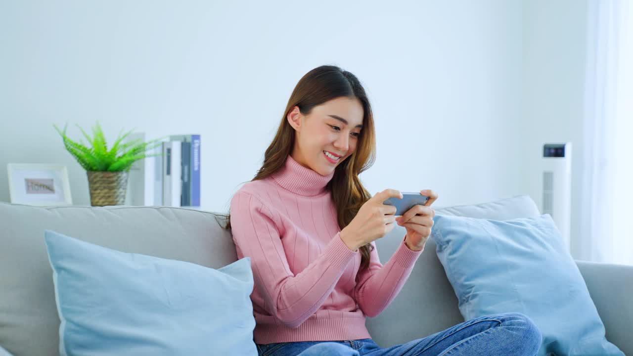 亚洲年轻美女在家里用智能手机玩手机游戏。迷人休闲的女孩感到快乐和放松，坐在沙发上玩手机触摸屏玩视频在家里享受胜利视频素材