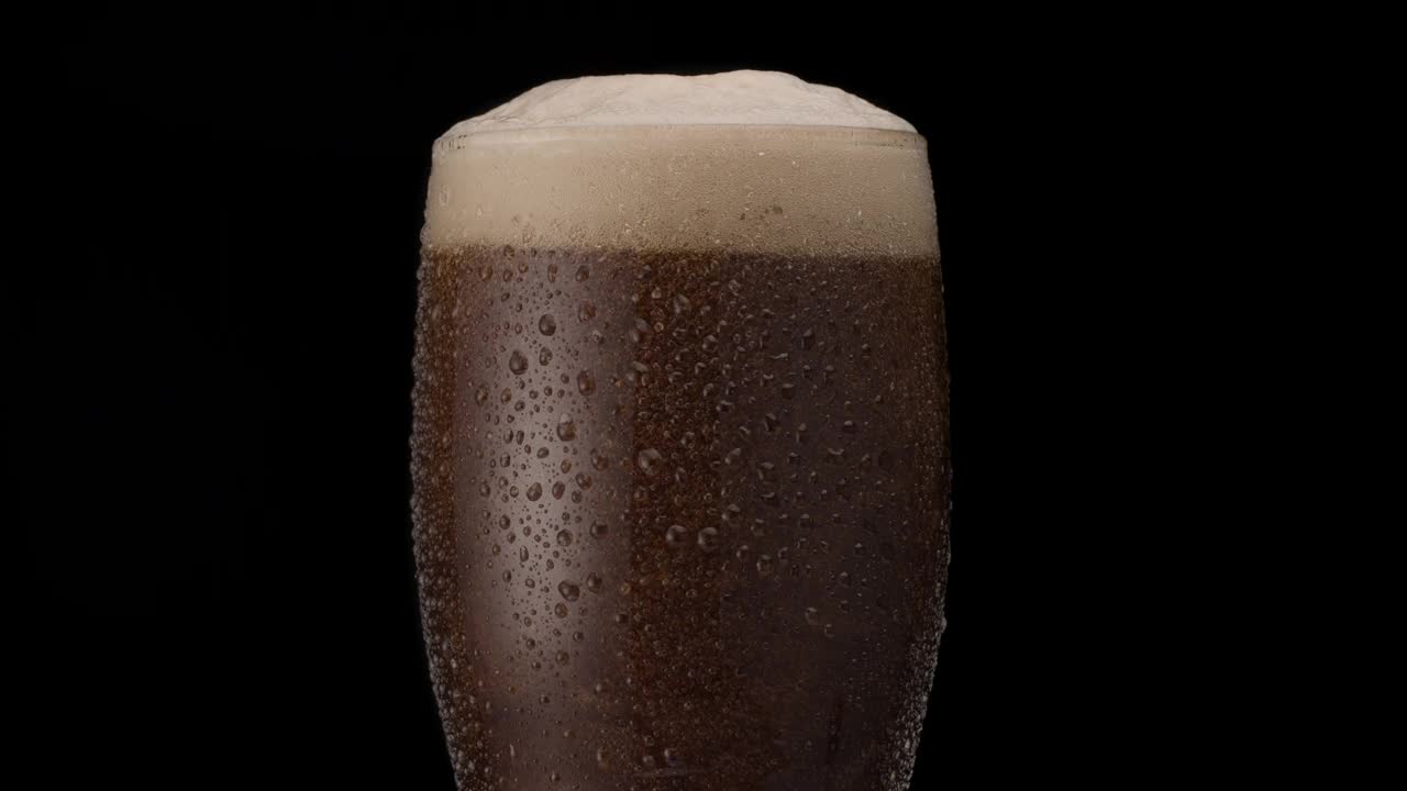 黑色背景上的黑色啤酒。啤酒在玻璃杯中晃动，泡沫和泡沫升起。一杯啤酒缓缓地顺时针旋转。视频素材