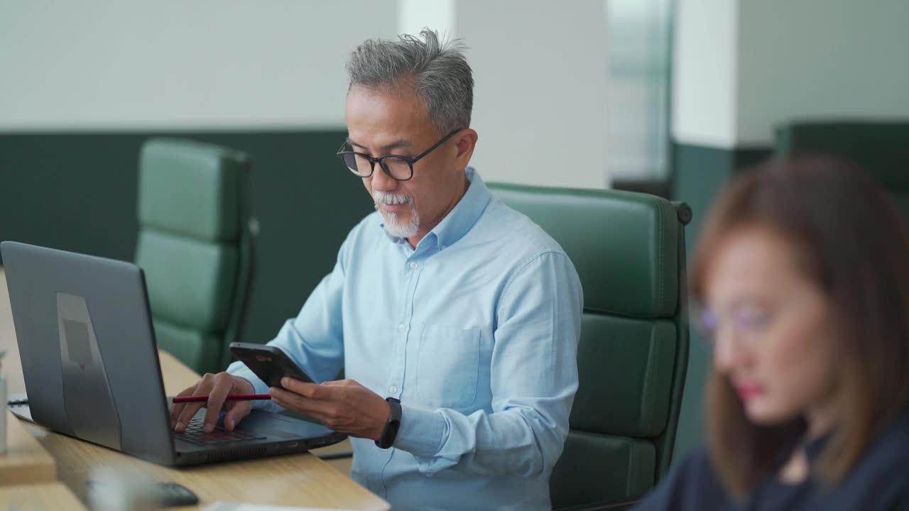 亚洲华人工作高级男子系使用笔记本电脑与他的同事在开放式办公室工作视频下载