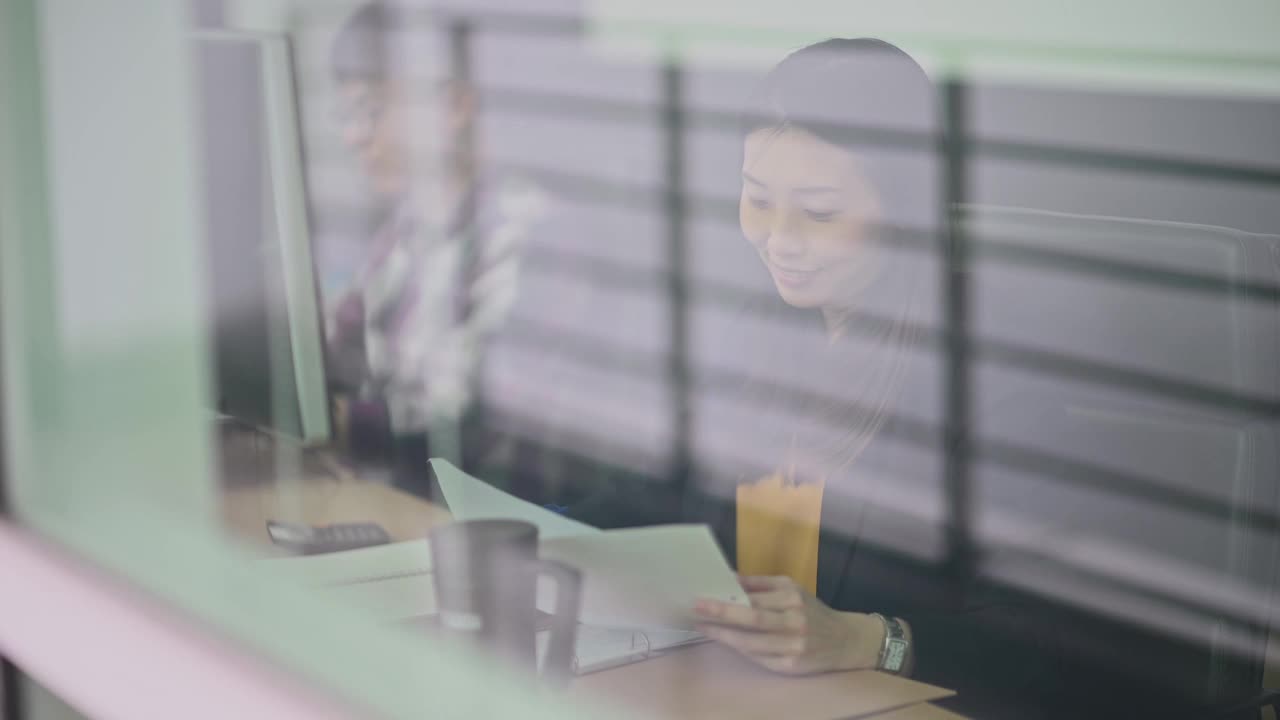 透过窗户，一位亚洲华人女性专注于阅读文献研究，与同事并肩工作在工作站视频素材