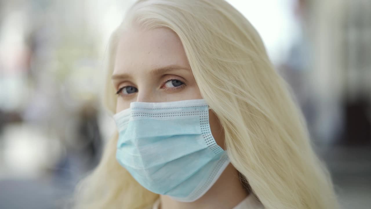 一个戴着医用口罩的蓝眼睛金发女人站在一个大城市的中心街道上看着摄像机。冠状病毒疫情、个人防护装备、检疫隔离。视频下载