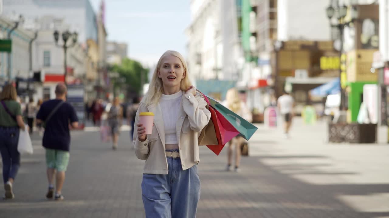 在大城市的精品店拿着包的时髦女人。她背着一堆彩色的袋子走在步行街上。她手里拿着一个原装的杯子，里面装着美味的咖啡。时装店和服装店。视频素材