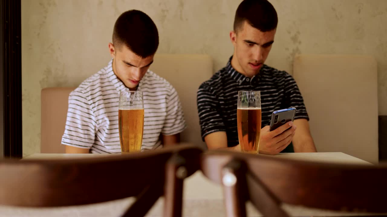 同卵双胞胎兄弟用啤酒敬酒，坐着和使用智能手机视频素材