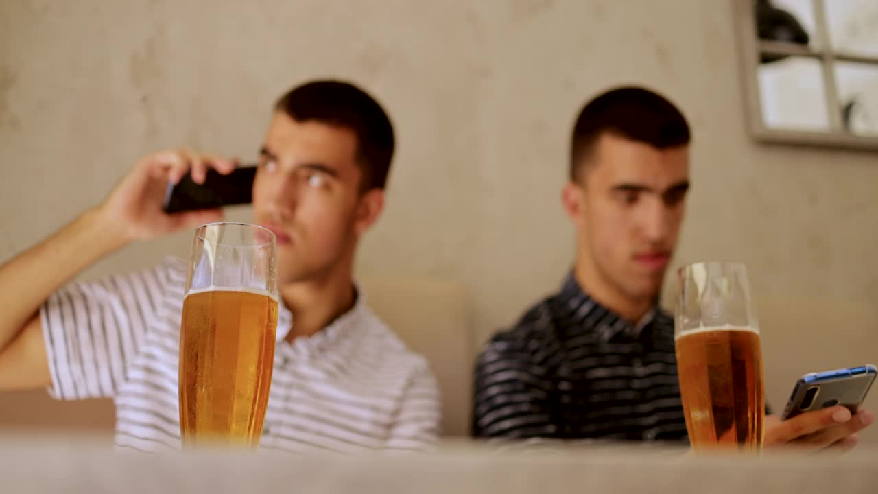 一模一样的双胞胎兄弟坐着，微笑着，喝着啤酒，用着智能手机视频素材