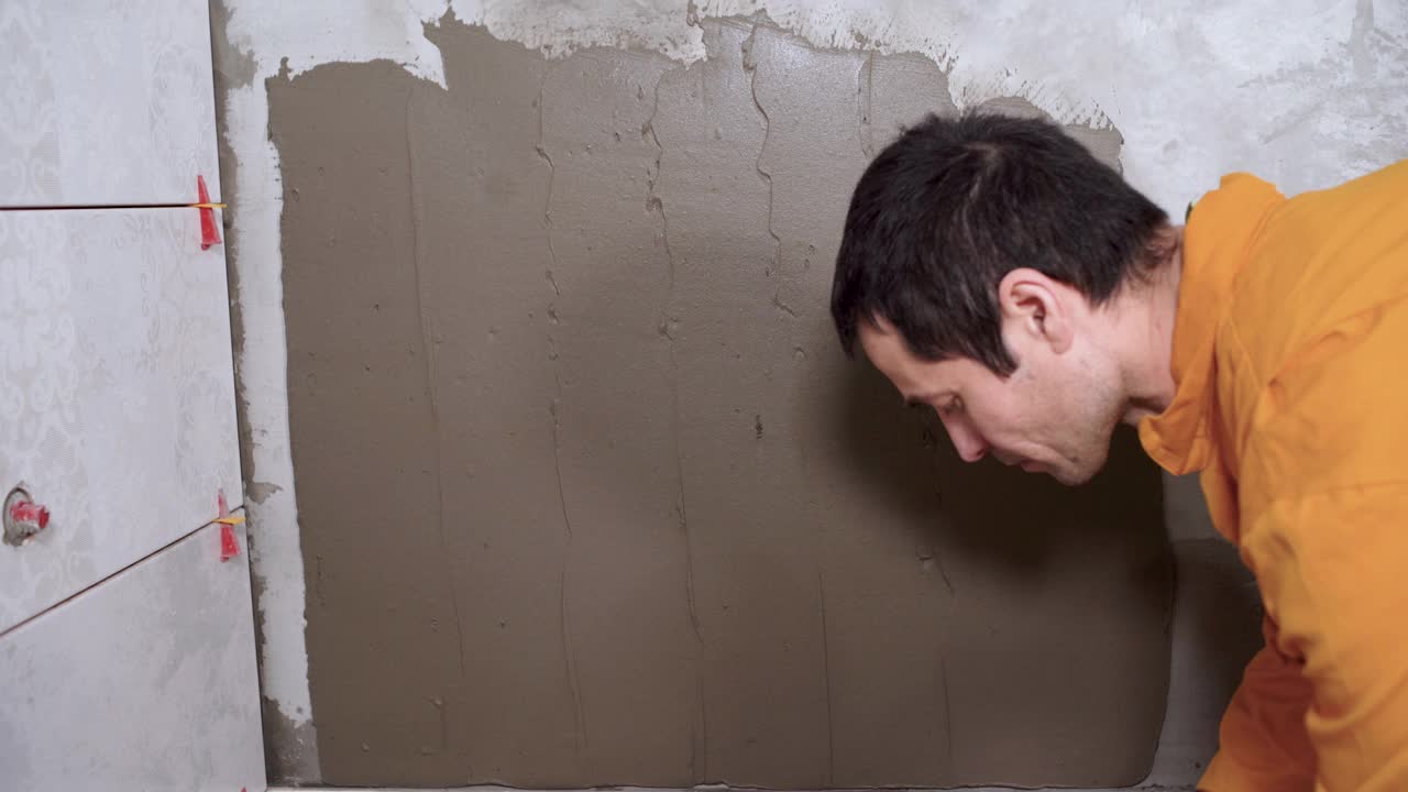 男建筑工用镘刀或抹刀在浴室墙上涂瓷砖粘合剂。瓷砖铺设工作的特写镜头。墙上做准备。改造的概念视频素材