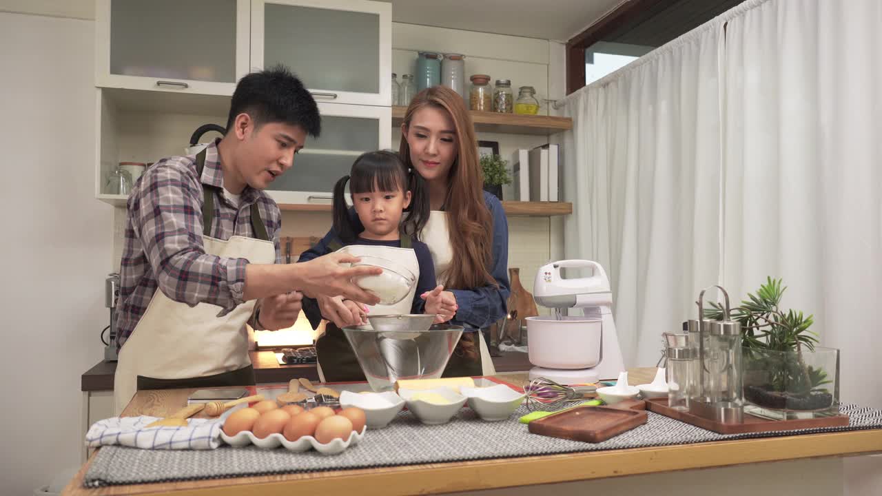 周末，亚洲父亲和母亲一起教日本女儿用面粉筛准备面粉。亚洲家庭的概念，一个孩子做饭，烘焙，烘焙。十几岁的女孩有积极的情绪和兴奋去做。视频下载