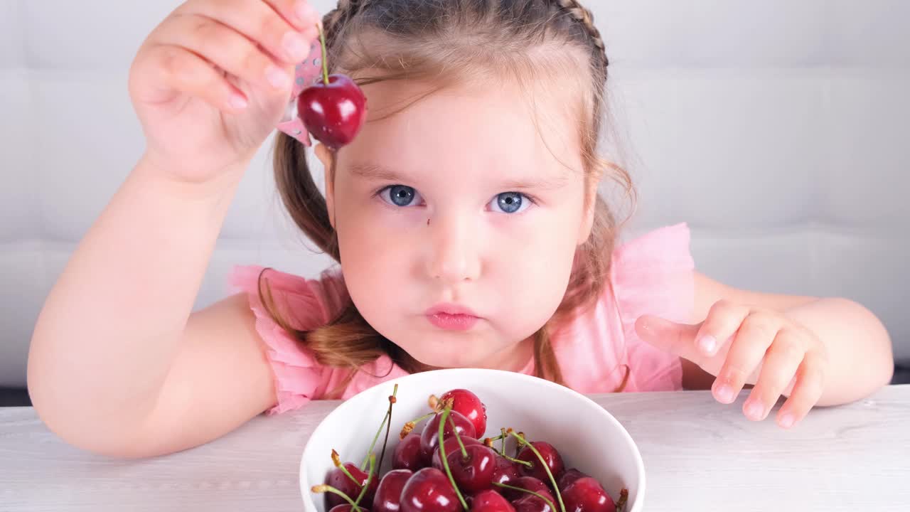 漂亮的小女孩坐在一张轻便的木桌旁，桌上放着一盘樱桃，手里拿着红色的浆果。健康饮食视频下载