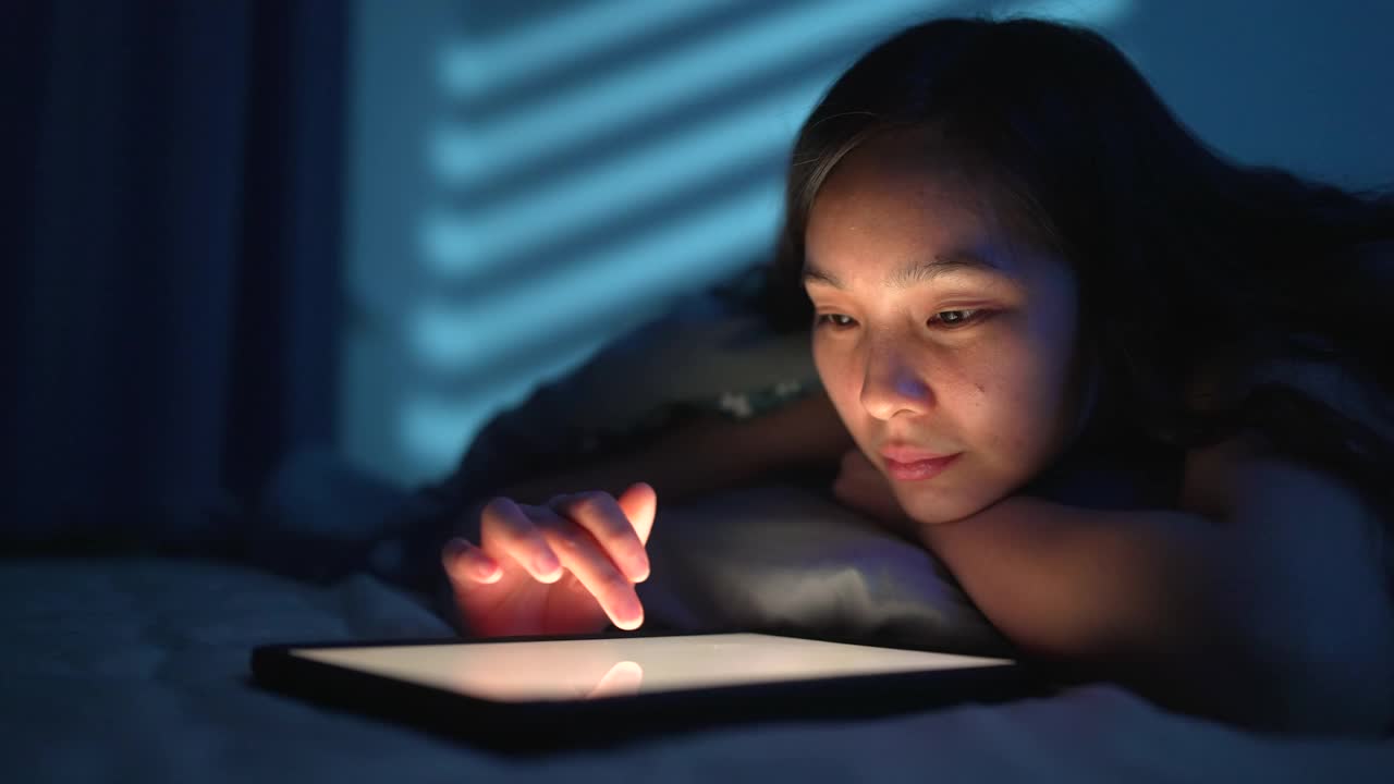 年轻的女孩面带微笑地躺在家里的床上玩着平板电脑视频素材