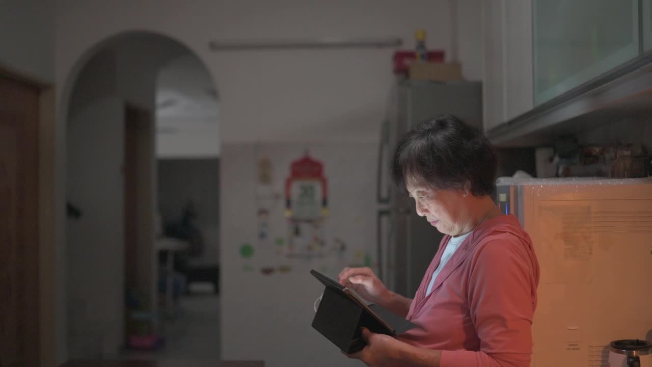 亚洲华人高级妇女在深夜使用数码平板电脑上网和在家庭厨房饮水视频素材