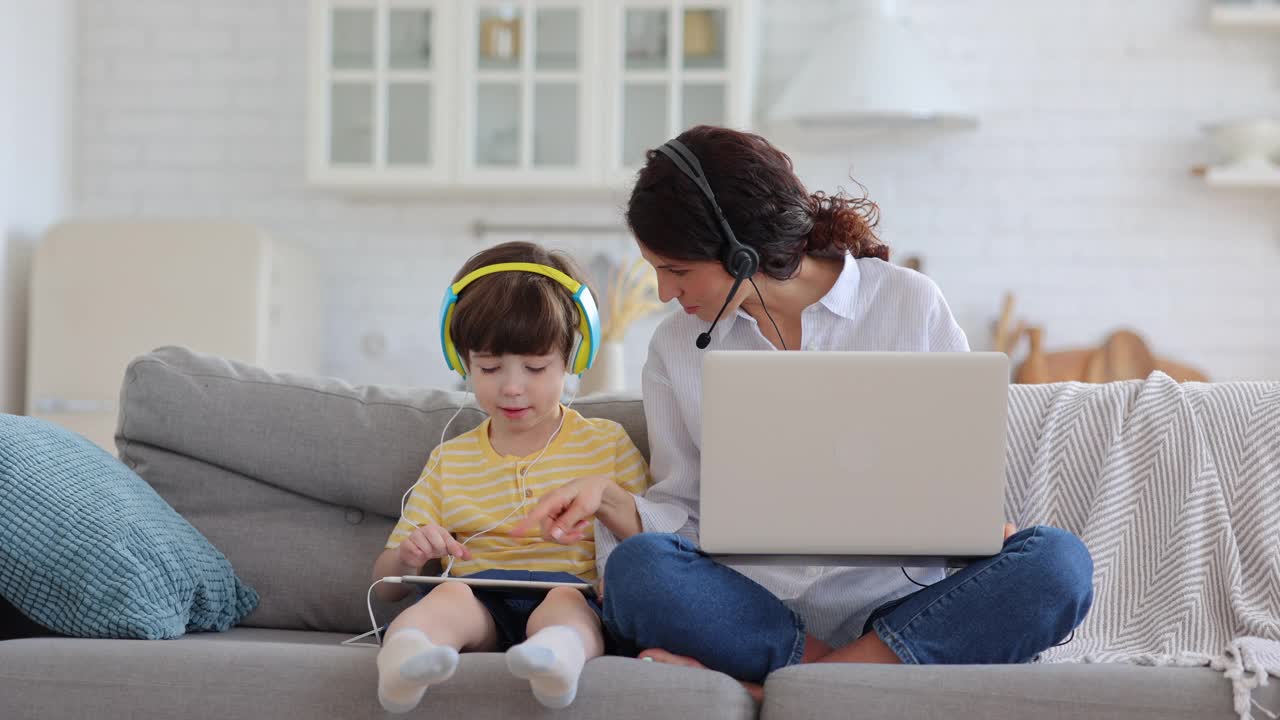 快乐的妈妈带着孩子坐在沙发上，在禁闭期间在笔记本电脑上工作，孩子在玩平板电脑视频素材