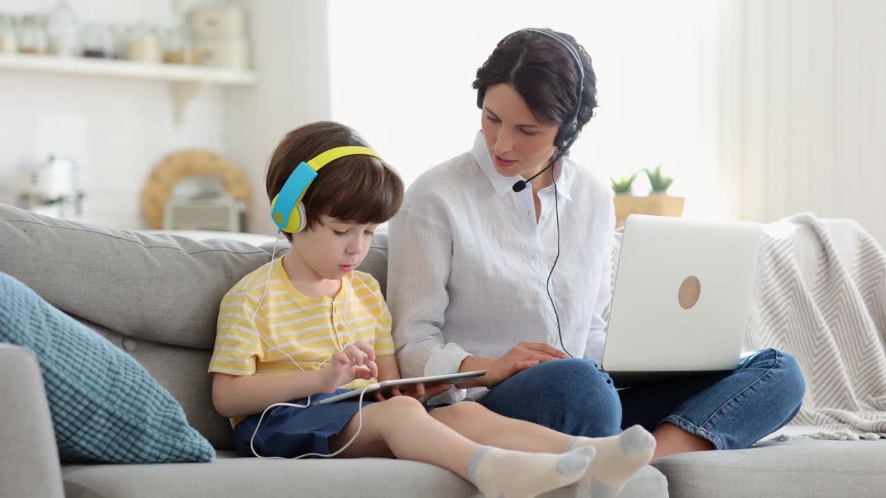 带着小孩在家工作:远程工作的妈妈给儿子数码平板电脑，用笔记本电脑开会视频素材