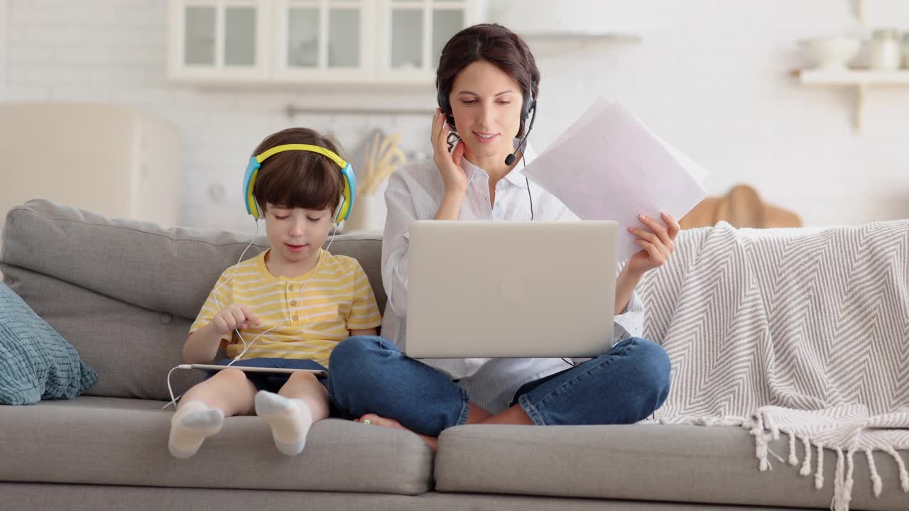 自由职业者妈妈在禁闭期间坐在沙发上办公，用笔记本电脑工作，孩子在玩平板电脑视频素材