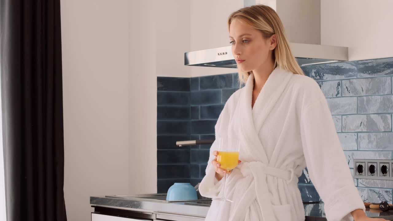 跟踪拍摄到一个穿着白色浴衣的漂亮金发女人在家里的厨房里愉快地喝着橙汁视频下载