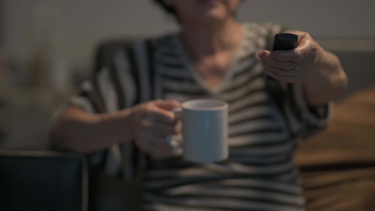 亚洲华人老年妇女手握电视遥控器，选择频道，在周末的闲暇时间独自一人在家看电视视频素材