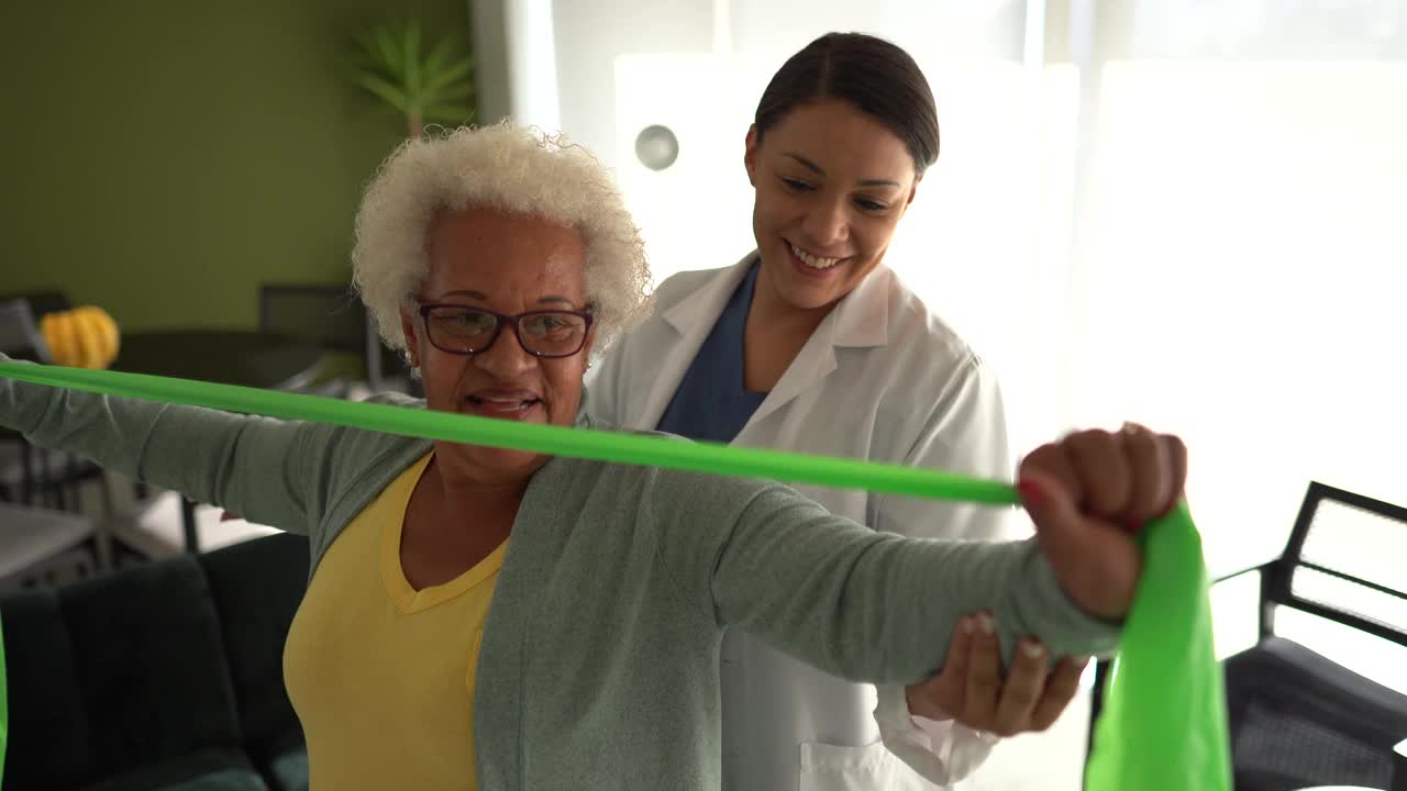 理疗师帮助老年妇女在家里用阻力带做练习视频下载