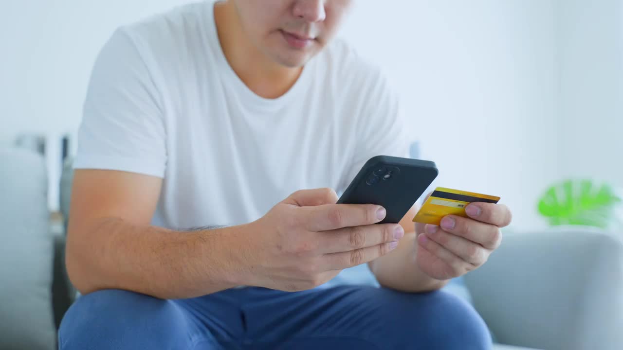 亚洲男性在家中客厅享受网上购物特写。帅哥坐在沙发上，用手机在店内网店购物后，拿着信用卡进行网上银行支付。视频素材