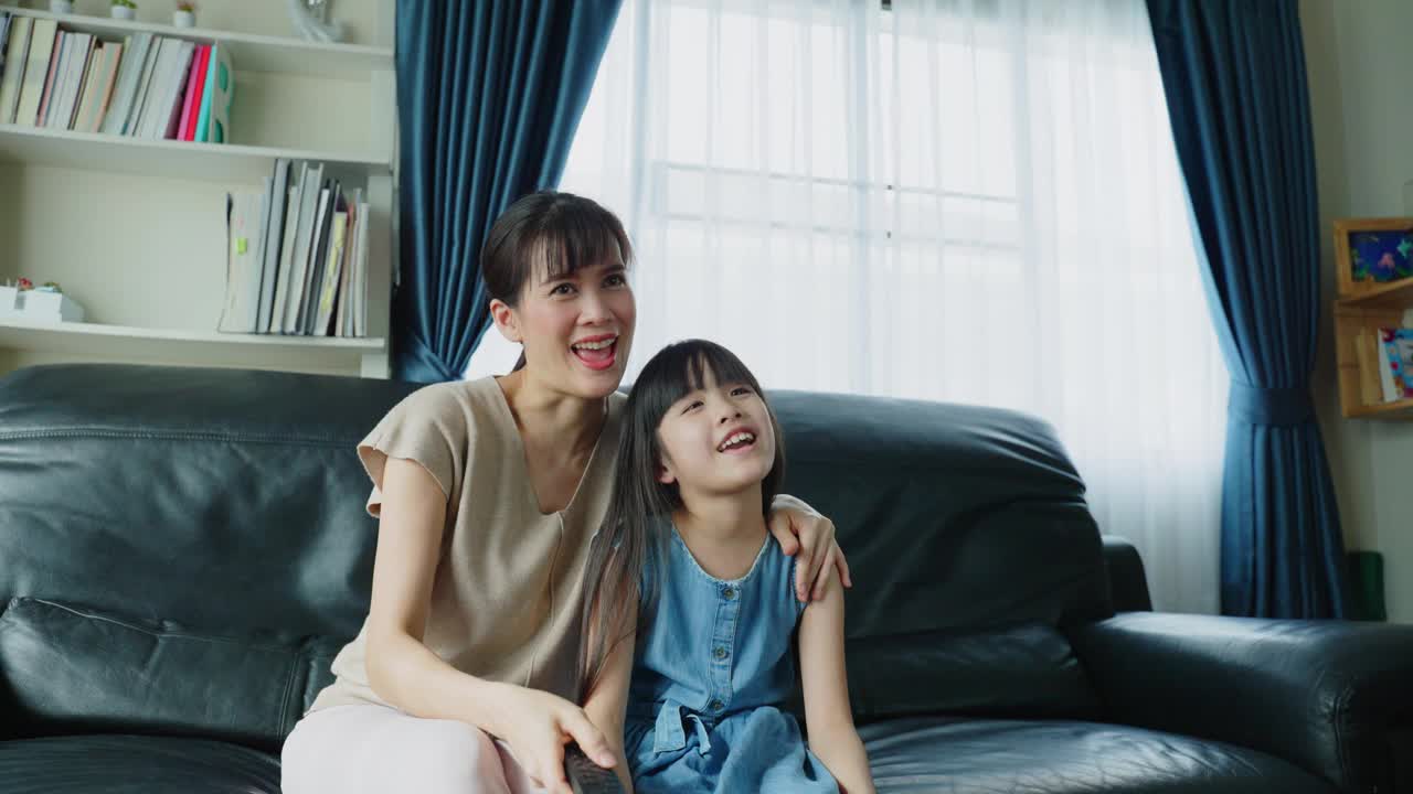 亚洲妈妈和孩子一起看电视上的喜剧电影，一起笑。幸福的家庭呆在家里，父母坐在沙发上，和小女儿在家里的客厅里度过闲暇时光。视频素材