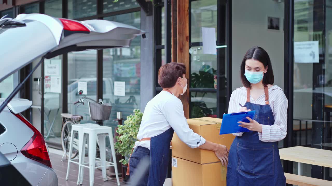 亚洲咖啡馆老板夫妇因新冠肺炎疫情戴口罩，在咖啡店工作。服务员把包裹装进车里，女招待的支票夹在写字板上。外购运输概念视频下载