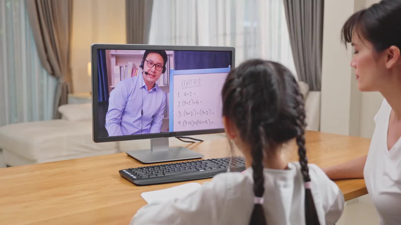 由于冠状病毒大流行，亚洲小女孩通过数字远程互联网会议向学校老师学习在线课程。孩子看着电脑，在妈妈的帮助下写纸条视频素材