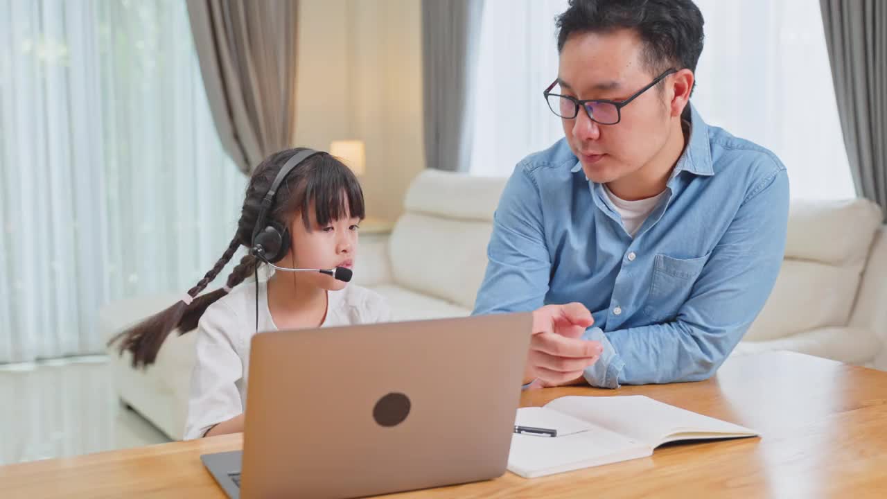 由于冠状病毒大流行，亚洲小女孩通过数字远程互联网会议向学校老师学习在线课程。孩子看着电脑，在爸爸的帮助下写纸条视频素材