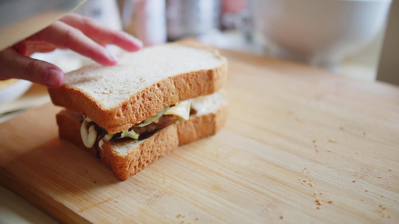 近距离的人在厨房的木板上用锋利的刀把三明治面包和肉、奶酪和蔬菜切成两半，在家做快餐视频下载
