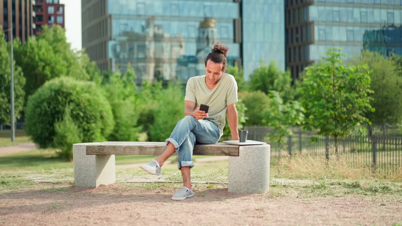 放大一个年轻人在工作休息时坐在公园的长椅上，一边用手机发短信一边思考，旁边放着笔记本电脑和外卖咖啡杯视频素材