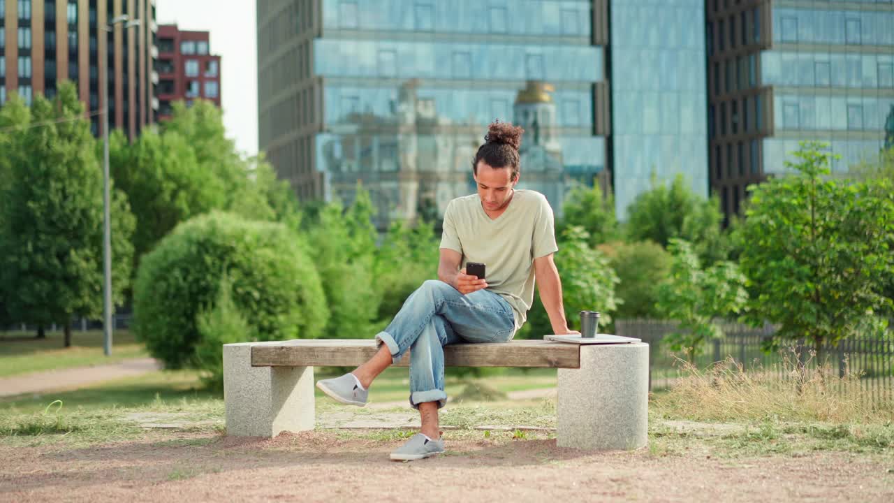 一头扎着马尾辫的年轻男子坐在公园的长椅上，一边用手机发短信，一边想着或做着白日梦，旁边放着笔记本电脑和外卖咖啡杯视频素材