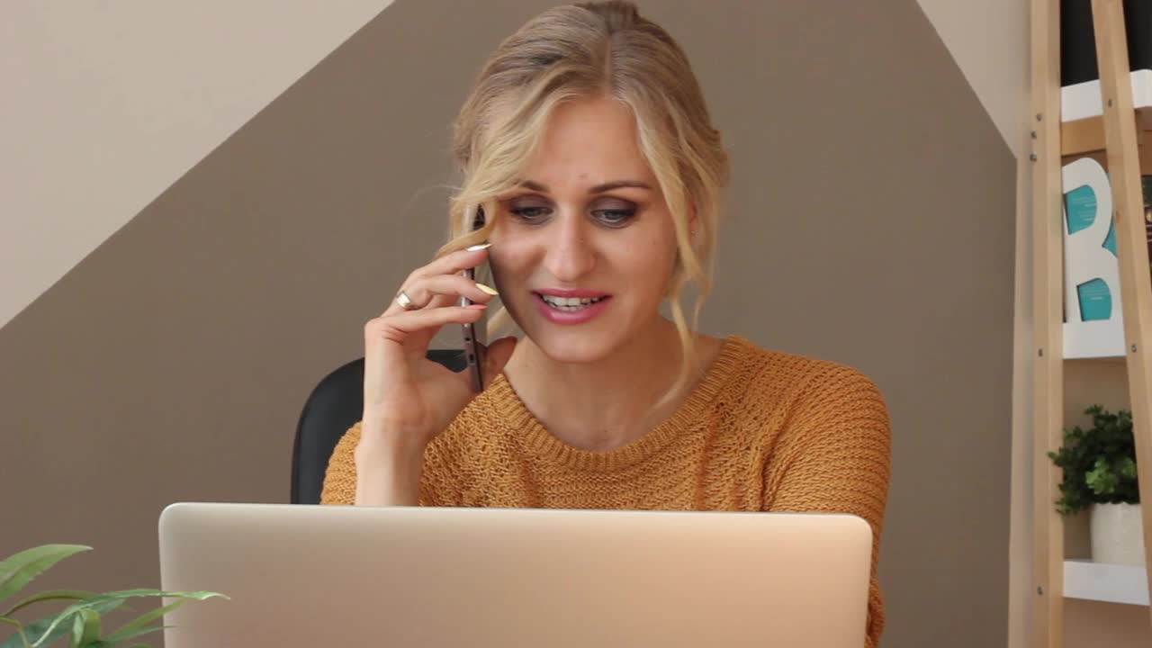 穿着居家衣服的女人坐在笔记本电脑前工作，讲着电话，动情地打着手势，甜甜地笑着视频素材