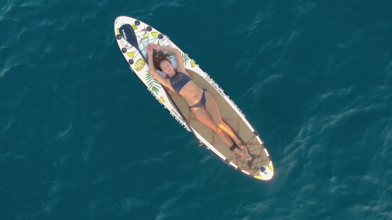 女人在冲浪板上漂浮。美丽的晒黑的女人躺在她的背部在比基尼和游泳平稳在小波浪。旁边放着一把桨。海是蓝色的。温暖的天气。夏天视频下载