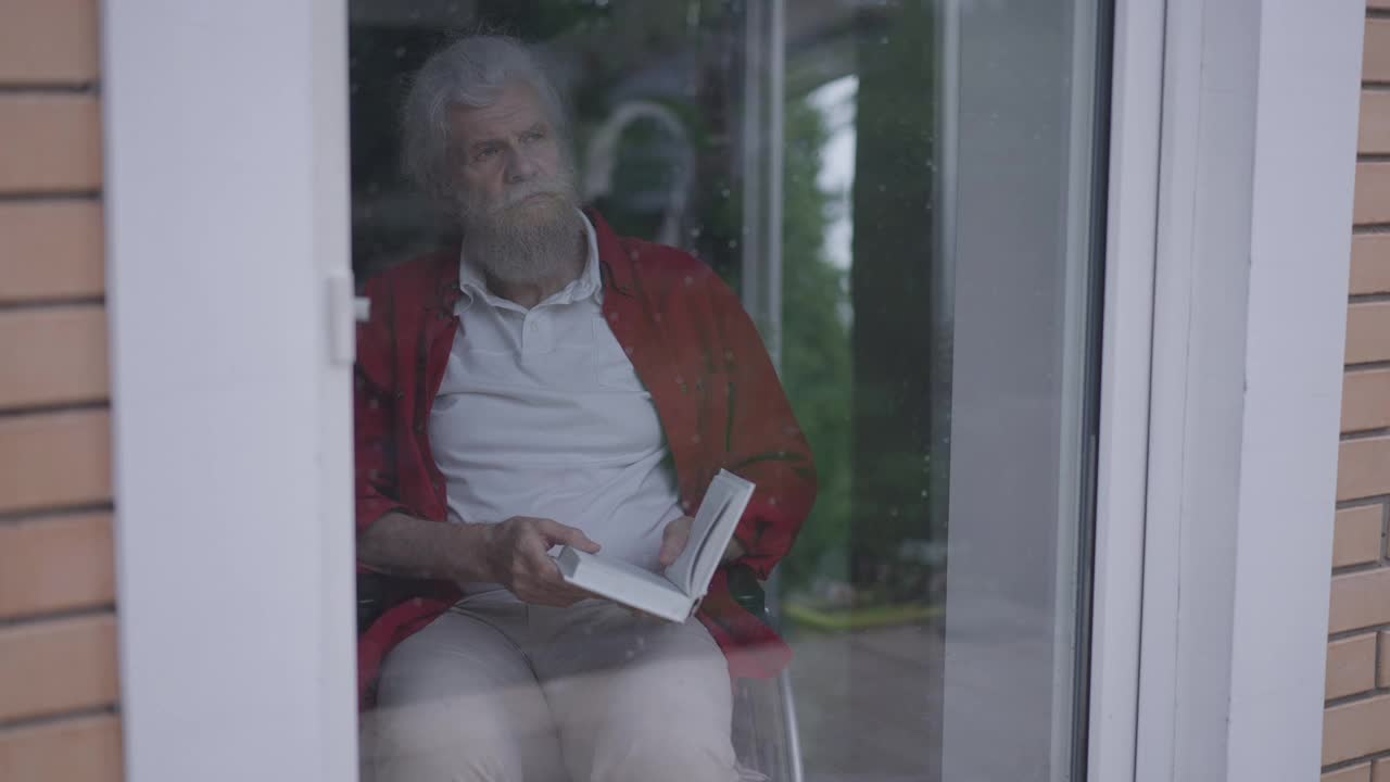 沮丧、深思熟虑的老年残疾人合上书，望着玻璃门上的窗外。沮丧孤独的白种人胡子残疾退休人员的肖像在室内。缓慢的运动。视频素材