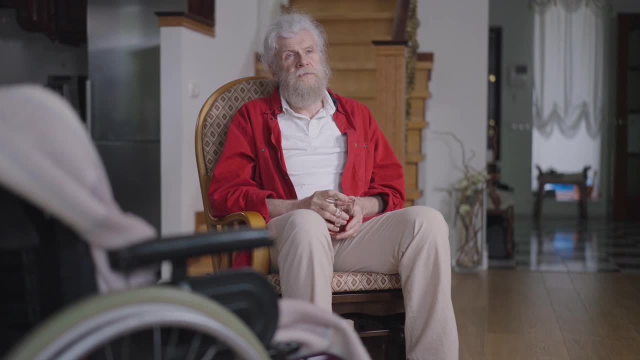 自信英俊的男子坐在舒适的扶手椅上喝茶，前面坐着模糊的轮椅。胡子灰白头发的白人残疾退休在家室内客厅。视频素材