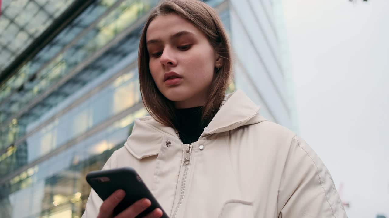 一个穿着羽绒服的休闲女孩的肖像，带着手机，若有所思地守候在城市的街道上视频素材