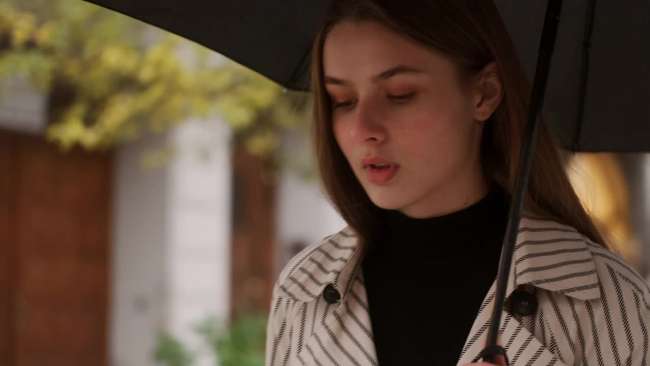 美丽时尚的女孩的肖像，穿着风衣，撑着伞走在下雨的城市街道视频素材