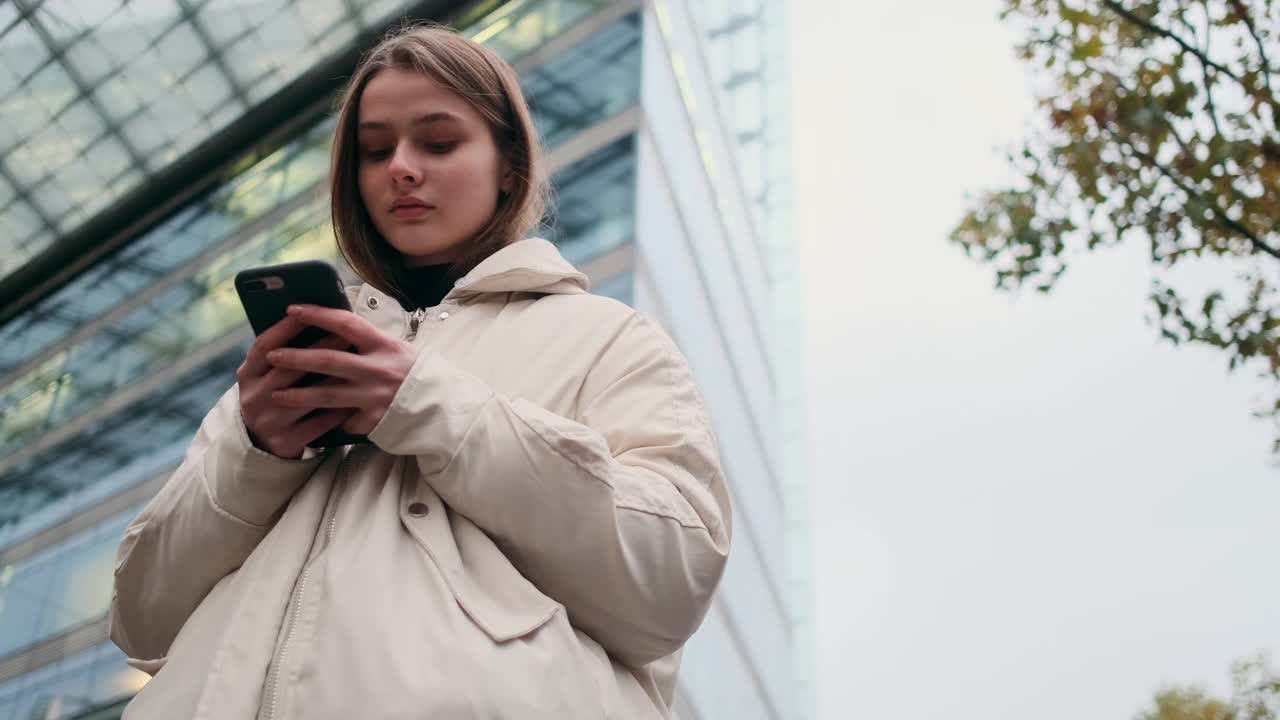 穿着羽绒服的休闲女孩在城市街道上愉快地使用手机等待视频素材