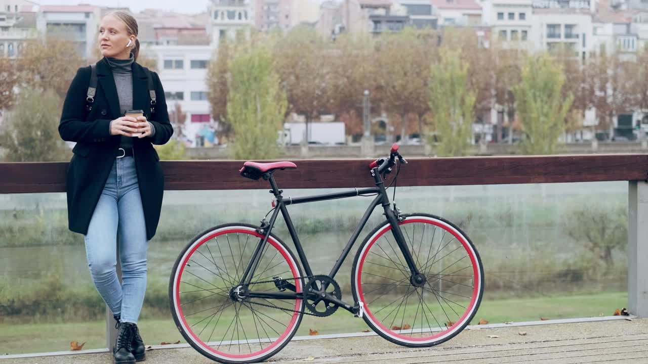 一个女人骑着自行车站在城市里，戴着耳机，拿着咖啡杯视频素材