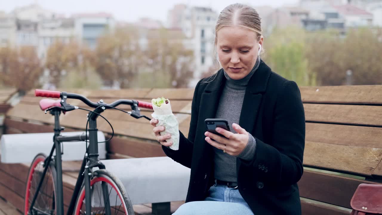 一位女士坐在长凳上，拿着包装好的三明治，用着智能手机视频素材