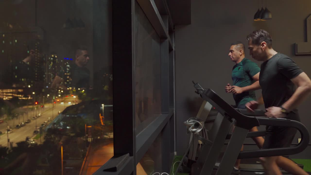 侧视宽镜头身体强壮的男人一起在健身房跑步机在晚上面对城市的街灯视频下载