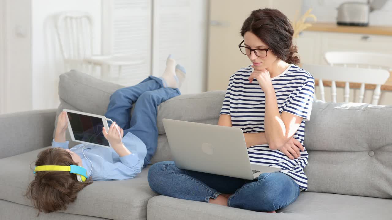 女人下班后坐在沙发上用笔记本电脑休息，孩子在玩平板电脑。封锁。视频素材