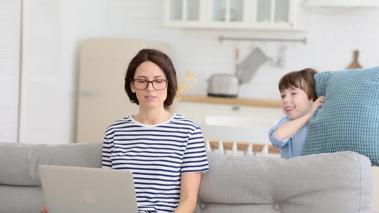 疲惫的母亲坐在家里的沙发上用笔记本电脑工作，孩子分散了注意力，用枕头攻击母亲视频素材