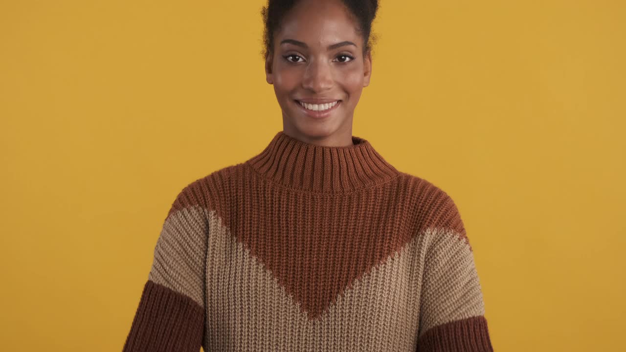 欢快的非洲裔美国女孩的肖像在针织毛衣愉快地在照相机在黄色的背景视频下载