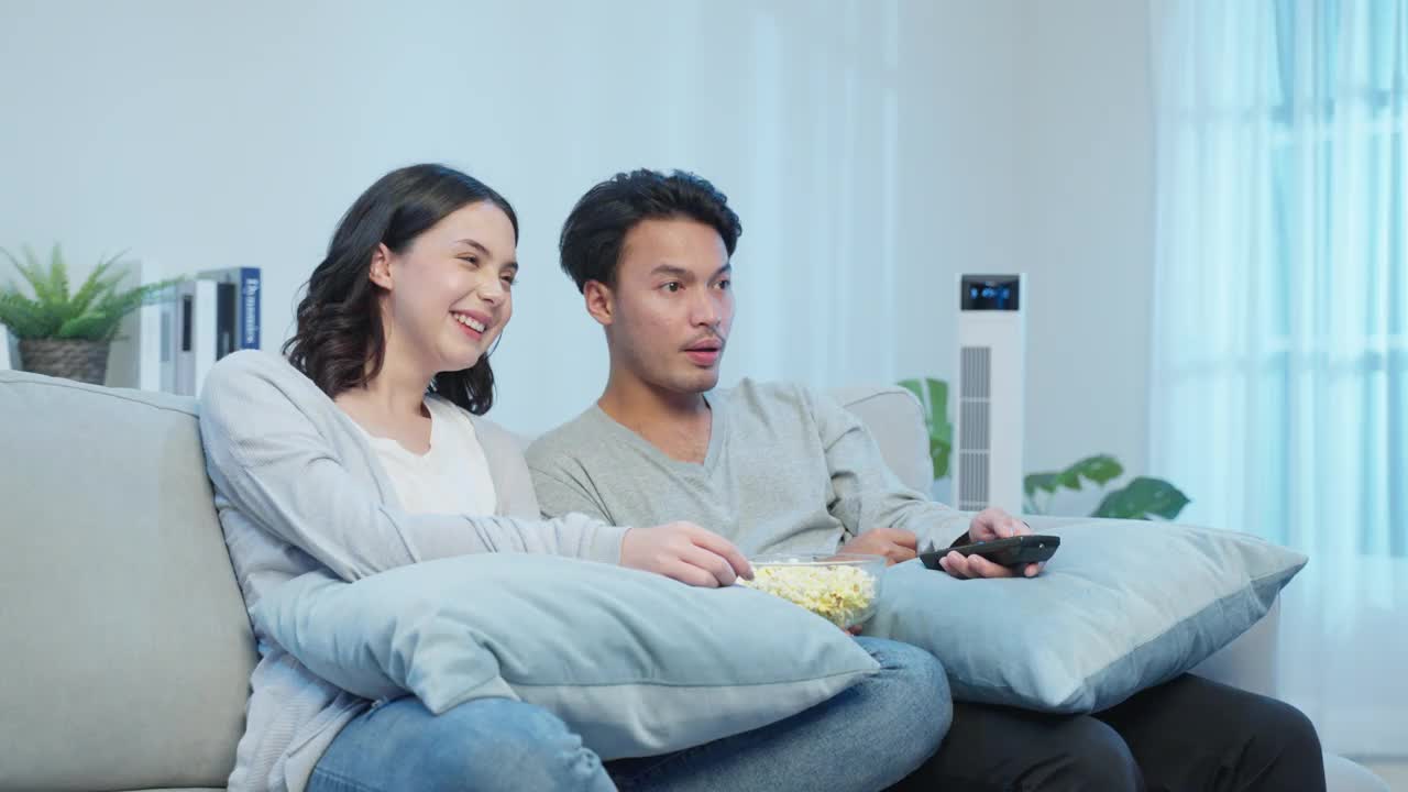 亚洲年轻情侣在家里的客厅里一起看电影。漂亮的男女坐在沙发上，快乐的男友用遥控器看电视节目，和女友在家里开怀大笑视频素材
