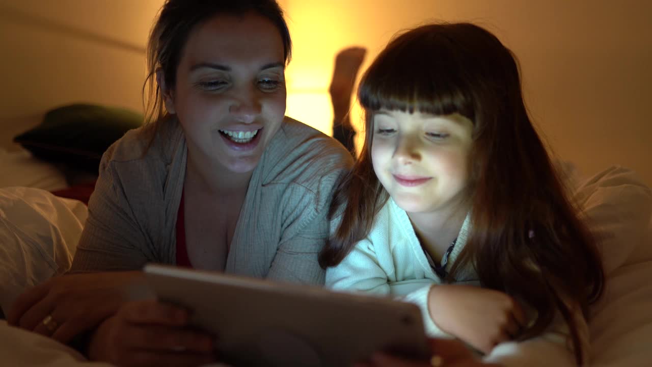 母亲和女儿躺在家里的床上用电子平板电脑看东西视频下载